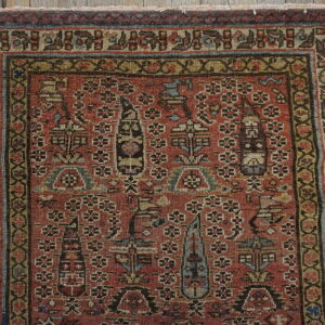Muted red rug featuring complex stylized motifs and elaborate borders, placed on distressed wooden flooring.