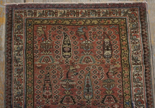 Muted red rug featuring complex stylized motifs and elaborate borders, placed on distressed wooden flooring.