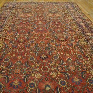 Rust-red rug featuring an intricate all-over stylized floral pattern and geometric borders on plank wood floors.