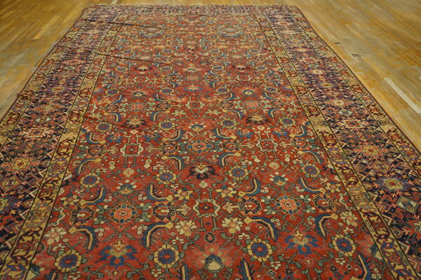 Rust-red rug featuring an intricate all-over stylized floral pattern and geometric borders on plank wood floors.