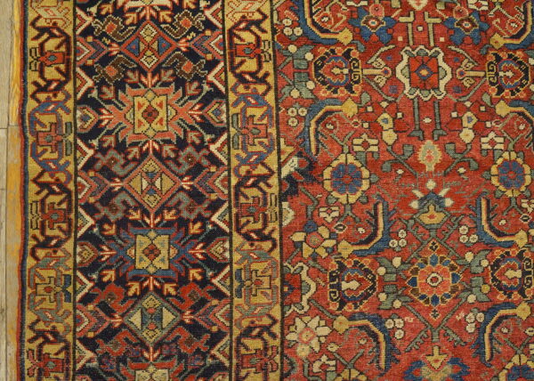 Densely woven rug detail shows geometric red and navy field motifs bordering a gold geometric pattern.