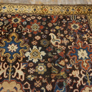 Detailed dark rug with colorful geometric and floral motifs, wide gold border, placed on wooden planks.