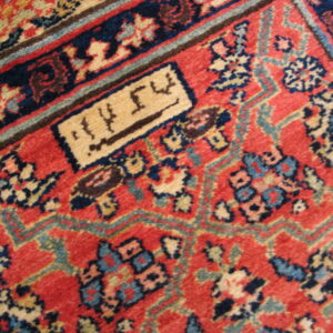 Close-up of a red patterned rug with navy, beige, and blue accents, featuring a rectangular cartouche with script.