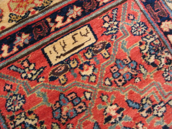 Close-up of a red patterned rug with navy, beige, and blue accents, featuring a rectangular cartouche with script.