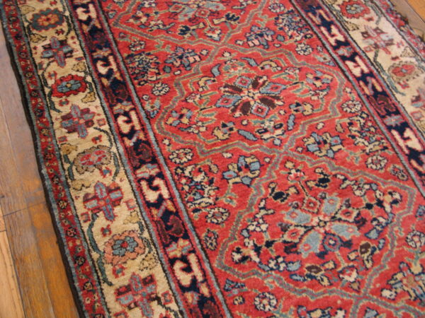 A traditional low-pile rug with red, navy, and beige intricate patterns rests on wood flooring.