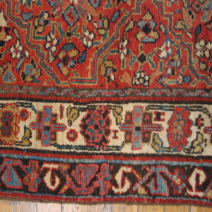 Close-up of a low-pile rug featuring a red field, blue and cream patterns, and detailed borders over wood flooring.