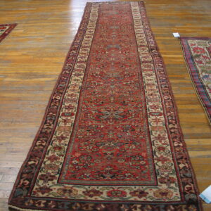 Long red runner rug with dense pattern on wood floor, visible next to two other traditional rugs.