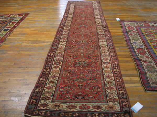 Long red runner rug with dense pattern on wood floor, visible next to two other traditional rugs.