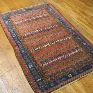 Low-pile rug with a red field, repeating colorful motifs, and wide blue borders on shiny hardwood planks.