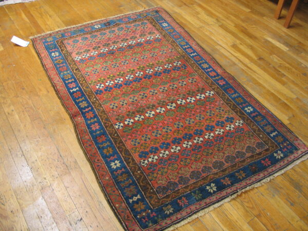 Low-pile rug with a red field, repeating colorful motifs, and wide blue borders on shiny hardwood planks.