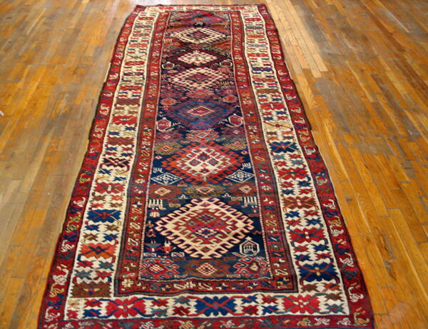 Geometric runner rug with stylized medallions and complex borders in red, blue, and off-white on wood floor.