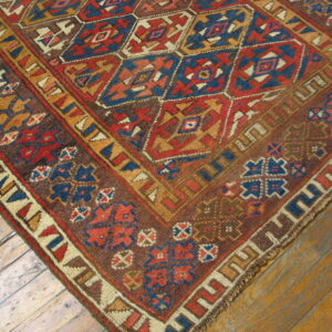 Low pile rug with complex red, blue, and gold geometric patterns rests on wood planks.