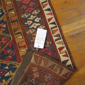 A folded rug with geometric patterns in red, navy, brown, and tan sits on honey-toned wood flooring.