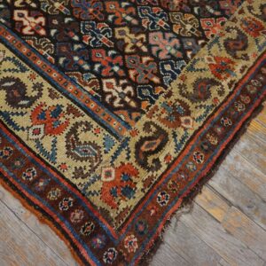 Geometric rug corner featuring rust, blue, tan, and brown colors, resting on distressed gray and yellow hardwood.