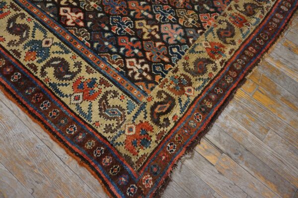 Geometric rug corner featuring rust, blue, tan, and brown colors, resting on distressed gray and yellow hardwood.