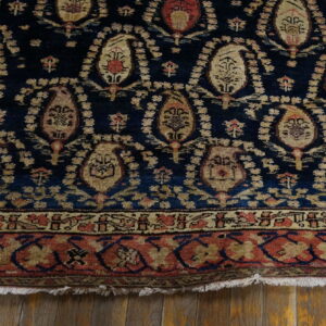 Navy blue rug with distressed tan and red paisley patterns and complex borders over light wood planks.