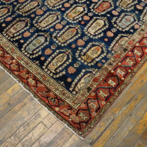 Blue rug corner featuring ogee motifs and a geometric red border resting on distressed, weathered hardwood flooring.