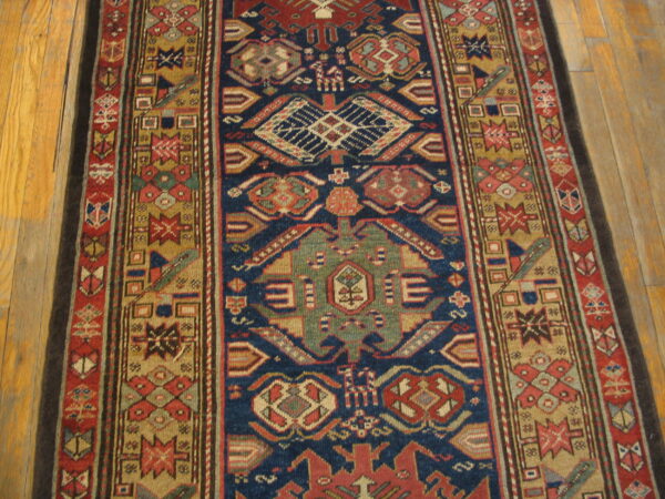 Traditional runner rug with complex geometric motifs in navy blue, rust, green, and tan on wood planks.