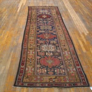Low-pile geometric runner rug with a navy, red, and gold pattern, displayed on worn wood planks.