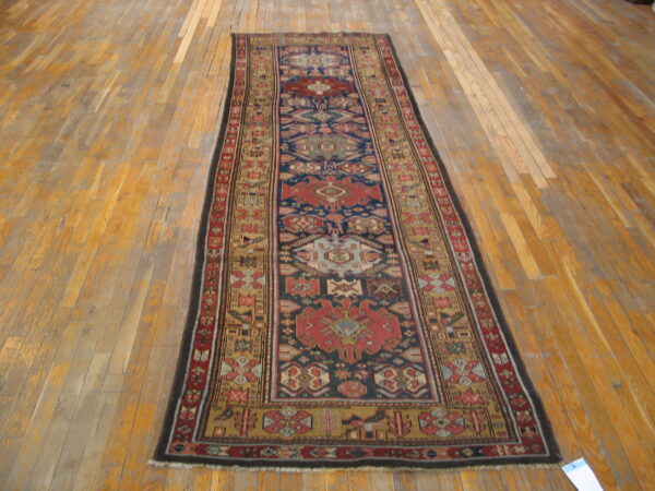 Low-pile geometric runner rug with a navy, red, and gold pattern, displayed on worn wood planks.