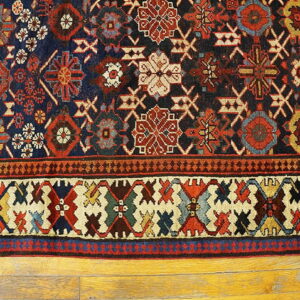A traditional rug with complex navy, red, and cream geometric patterns rests on yellow wood flooring.