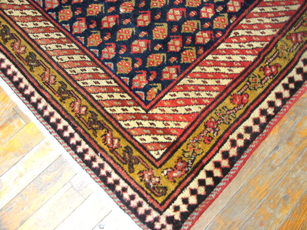 Navy, red, and gold geometric rug corner with striped borders rests upon light, worn wooden floorboards.