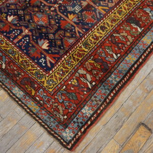 Intricate red, navy, and gold patterned rug corner with geometric borders sits on rustic gray wood floors.