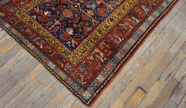 Intricate red, navy, and gold patterned rug corner with geometric borders sits on rustic gray wood floors.