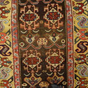 Rug close-up showing a dark brown field with red geometric motifs and wide gold borders on wood planks.