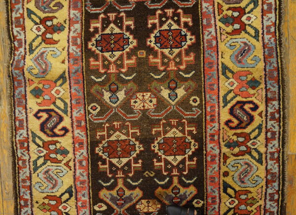 Rug close-up showing a dark brown field with red geometric motifs and wide gold borders on wood planks.