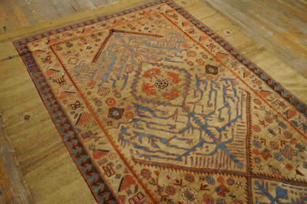 Rare collectable tribal camel color late 19th century antique nw persian serab rug 2 Geometric tan rug with stylized blue and rust patterns lies on a muted tan surface and distressed wood floor.