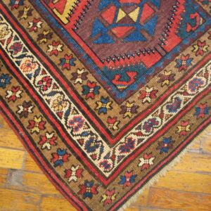 Geometric rug corner in deep red, blue, yellow, and brown lying on light hardwood flooring.