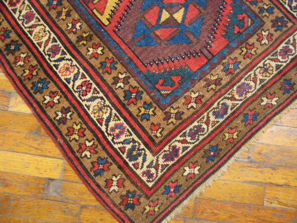 Geometric rug corner in deep red, blue, yellow, and brown lying on light hardwood flooring.