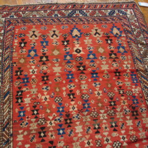 Red, low-pile rug with dense geometric patterns in blue, black, and tan resting on wood plank flooring.