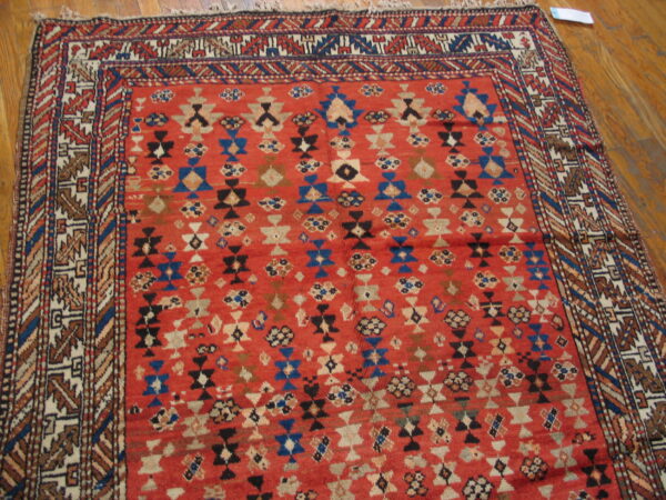 Red, low-pile rug with dense geometric patterns in blue, black, and tan resting on wood plank flooring.