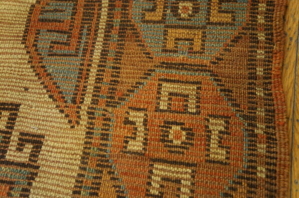 Geometric flatweave rug texture in rust, brown, and teal, resting beside a light wood floor.