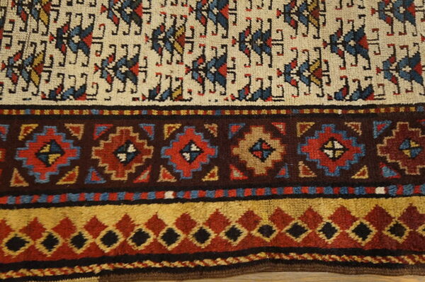 Textured rug detail showing cream field motifs and stepped borders in dark brown, red, blue, and gold.