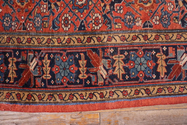 Detailed view of a low-pile rug in red, navy, and tan with complex borders on rustic wooden planks.