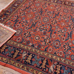 Ornate red and navy rug with fringe detail, partially flipped over on light wooden floorboards.