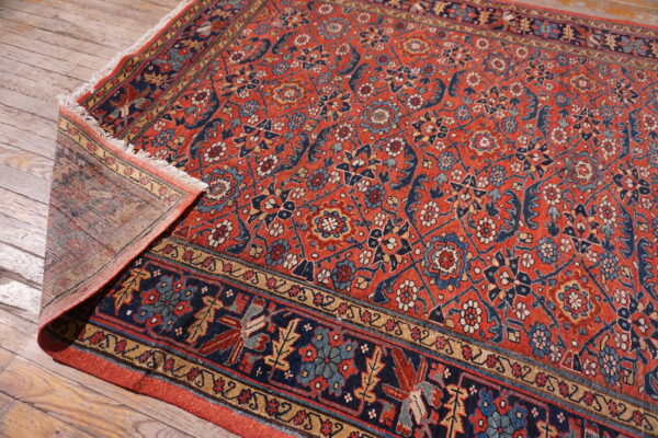 Ornate red and navy rug with fringe detail, partially flipped over on light wooden floorboards.