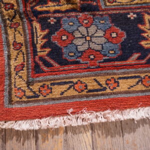 Low-pile rug detail showing red, blue, and gold geometric motifs resting on distressed wood.