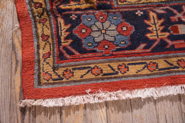 Low-pile rug detail showing red, blue, and gold geometric motifs resting on distressed wood.