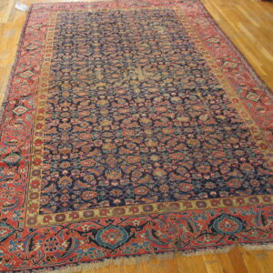 Navy blue area rug with a multicolor floral pattern and wide red borders rests on light hardwood flooring.