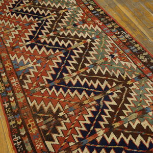Low-pile runner rug displays complex zigzag geometric patterns in rust, navy, brown, and green on wood floors.