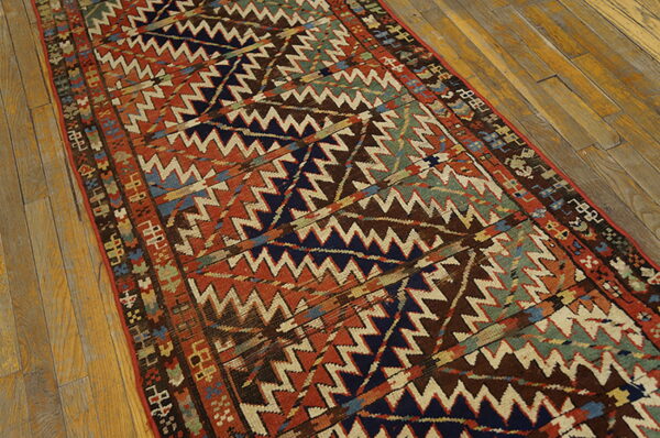 Low-pile runner rug displays complex zigzag geometric patterns in rust, navy, brown, and green on wood floors.