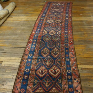 Geometric blue and rust runner rug displayed on worn wood floors with rolled textiles in the background.