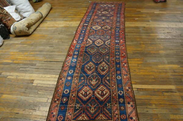 Geometric blue and rust runner rug displayed on worn wood floors with rolled textiles in the background.