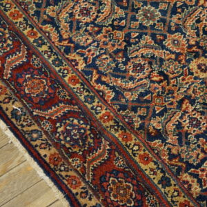 Close-up of a navy blue rug featuring dense, ornate patterns in red, tan, and gold, lying on wood planks.
