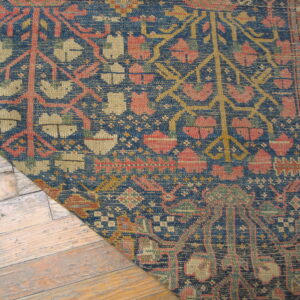 Low-pile rug with dense blue, coral, and khaki traditional pattern shown on light, worn wood flooring.