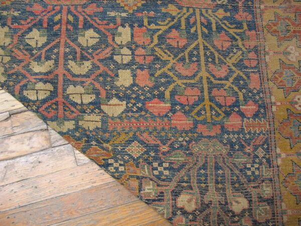 Low-pile rug with dense blue, coral, and khaki traditional pattern shown on light, worn wood flooring.